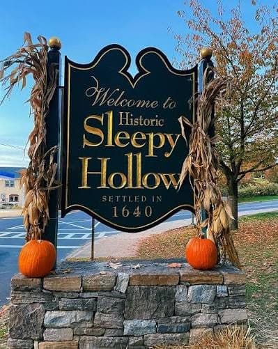A black and gold sign reading "Welcome to Historic Sleepy Hollow, Settled in 1640" on a stone base, flanked by corn stalks and two pumpkins.