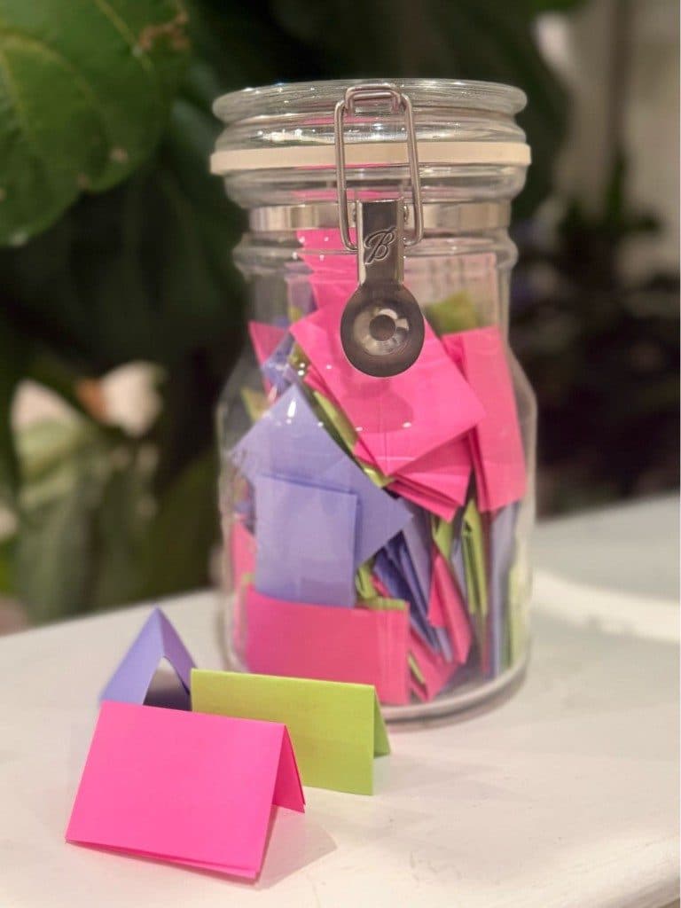 A glass clip-top jar packed with small folded notes in pink, purple and green, three of them set out on the white table in front of it